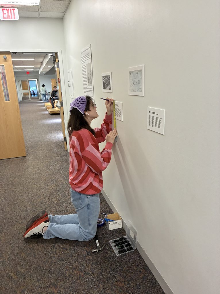 A person kneeling and measuring the wall with a ruler to hang artwork in a hallway, wearing a pink striped sweater and a purple headband.