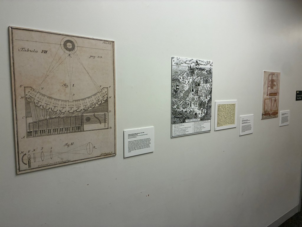 A collection of historical illustrations displayed on a wall, featuring a detailed scientific diagram, a map titled 'New Atlantic,' and other texts with descriptions.
