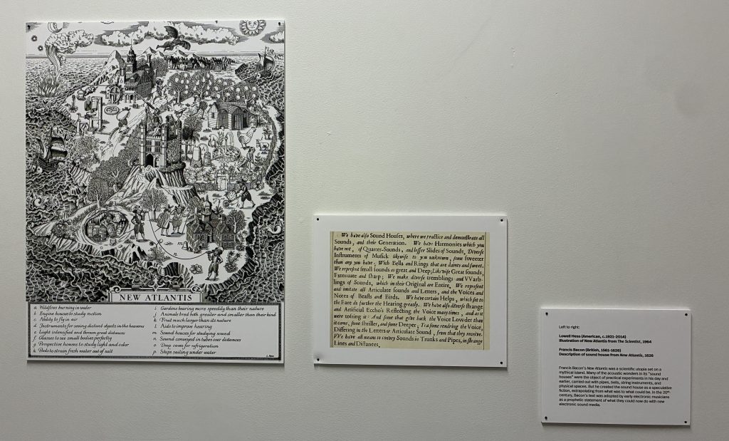 An artistic representation of 'New Atlantis', featuring various scenes of a mythical city with intricate buildings, landscapes, and people engaged in different activities, displayed on a wall alongside two informational plaques.